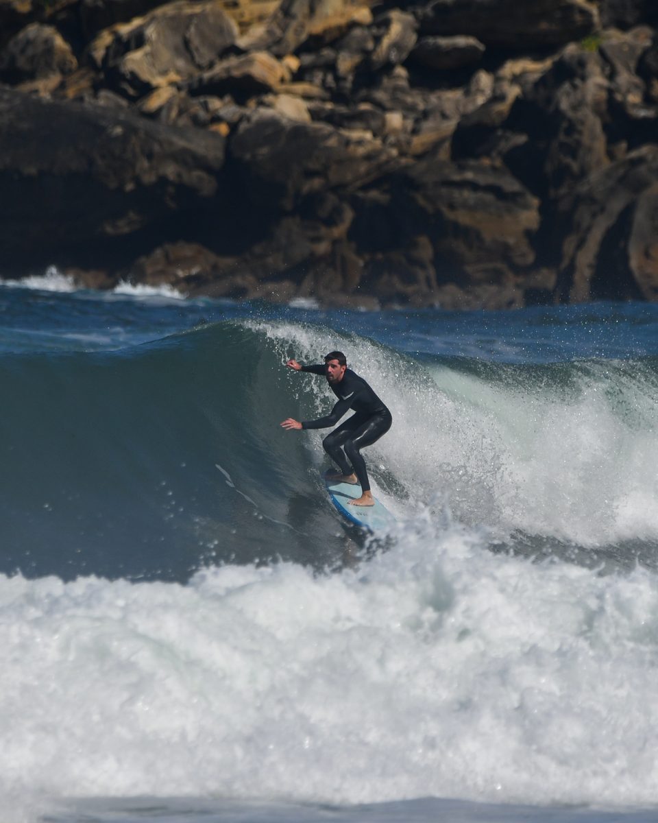 Un surfista sobre una ola en la Zurriola, con la costa rocosa al fondo.