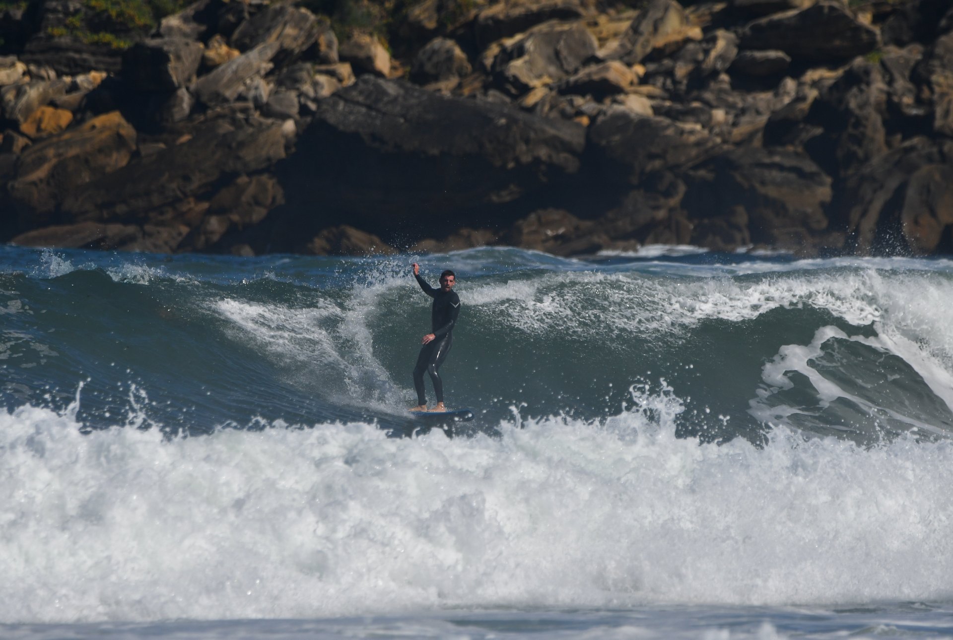 Un surfista levanta un brazo sobre una ola de la Zurriola, con las rocas de la costa al fondo.